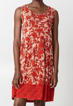 Robe sans manches rouge et beige à fleurs avec un ourlet rouge à motif portée par une personne debout devant un fond uni.