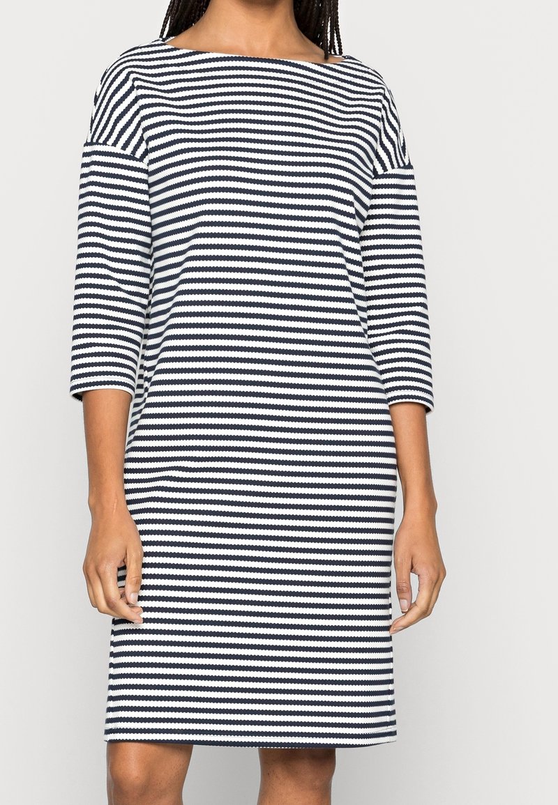 Femme portant une robe rayée horizontale noir et blanc longueur genou avec des manches trois-quarts et un décolleté bateau.