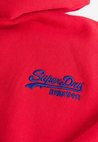 Červená tkanina s modrou vyšívanou logem "SuperDry" a japonskými znaky níže na složeném oděvu.
