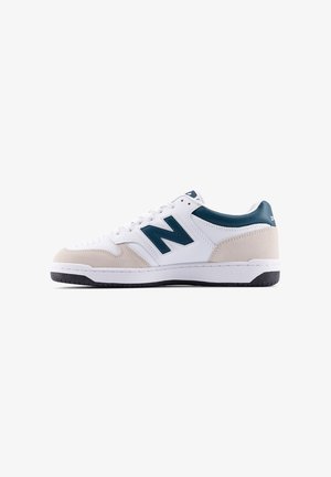 Zapatilla deportiva blanca con detalles en ante beige y logo en verde azulado oscuro. Cuenta con punta redondeada, cierre de cordones y suela de goma texturizada.