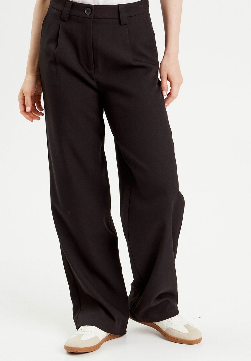 Pantalons à larges jambes en tissu noir, avec des plis à l'avant, des passants de ceinture et un fermeture à bouton. Associés à des baskets de couleur claire.