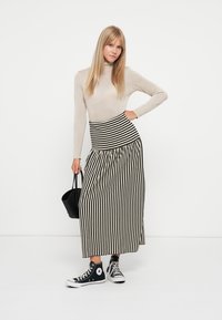 Beige langermet topp paret med et svart-hvitt stripet maxiskjørt. Modellen har på seg svarte joggesko og holder en svart håndveske.
