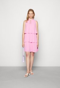 Vestido rosa em camadas com um decote atado, tecido texturizado e uma silhueta fluida. Combinado com sandálias brancas e uma pequena mala lavanda.