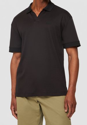 Mand iført en sort poloshirt med korte ærmer og blød krave samt beige bukser, stående mod en ensartet lys baggrund.