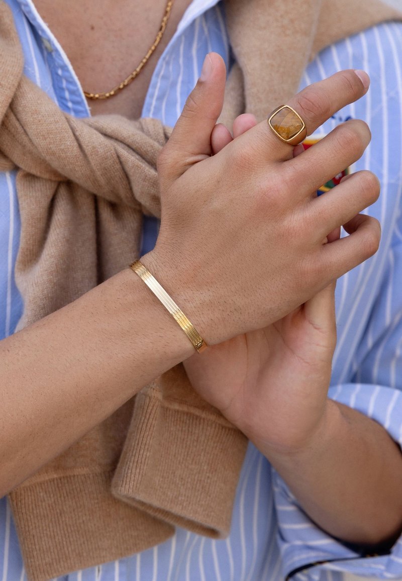 Gold metal bracelet with textured stripes and a square ring featuring a yellow gemstone. Hands resting with a beige sweater draped.