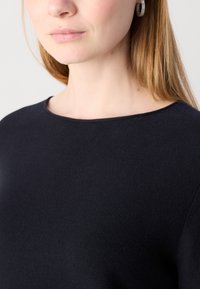 Črn pulover z teksturiranim zaključkom, okroglim vratnim izrezom in vidnimi ramenskimi šivi. Model ima dolge lase in nosi uhane v obliki obroča.