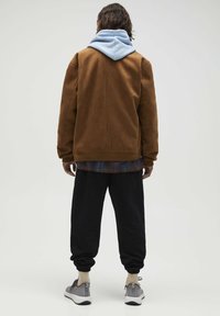 Bruine suede jas over een blauwe hoodie, gecombineerd met zwarte tapered broek en grijze sneakers. Zichtbare textuurdetails en geribbelde manchetten.