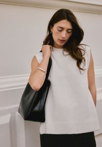 Haut en tricot blanc sans manches avec ourlet côtelé, porté avec un sac fourre-tout en cuir noir. Le mannequin porte un bracelet manchette argenté à son bras et a des cheveux ondulés et lâches.