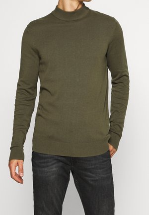 Mann trägt olivgrünen, gerippten Pullover mit Stehkragen und schwarze Jeans, steht mit einer Hand in der Tasche vor weißem Hintergrund.