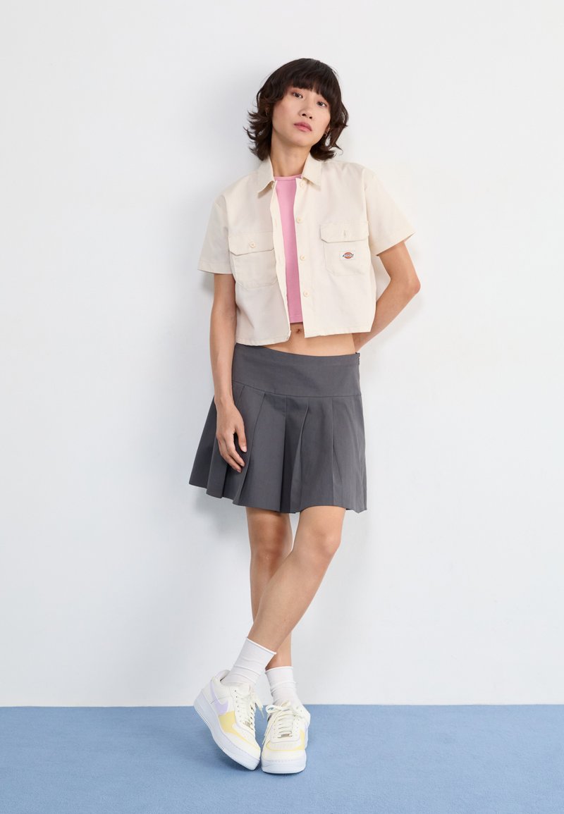 Camisa corta beige claro con dos bolsillos sobre una camiseta de tirantes rosa, combinada con una falda gris plisada y zapatillas blancas con acentos amarillos y morados.