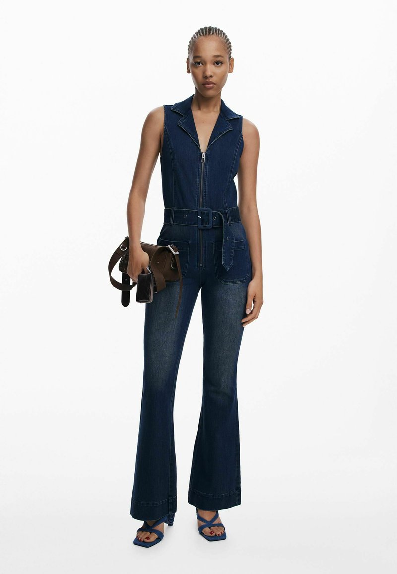 Jeansový overal so zipsom vpredu, golierom a pásom v páse. Rozšírené nohavice a hlboká modrá farba, doplnený malou hnedou taškou a modrými sandálmi.