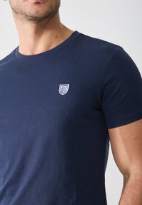 Tamnoplava pamučna majica s okruglim izrezom i kratkim rukavima, s malim bijelim izvezenim grbom na prsima.