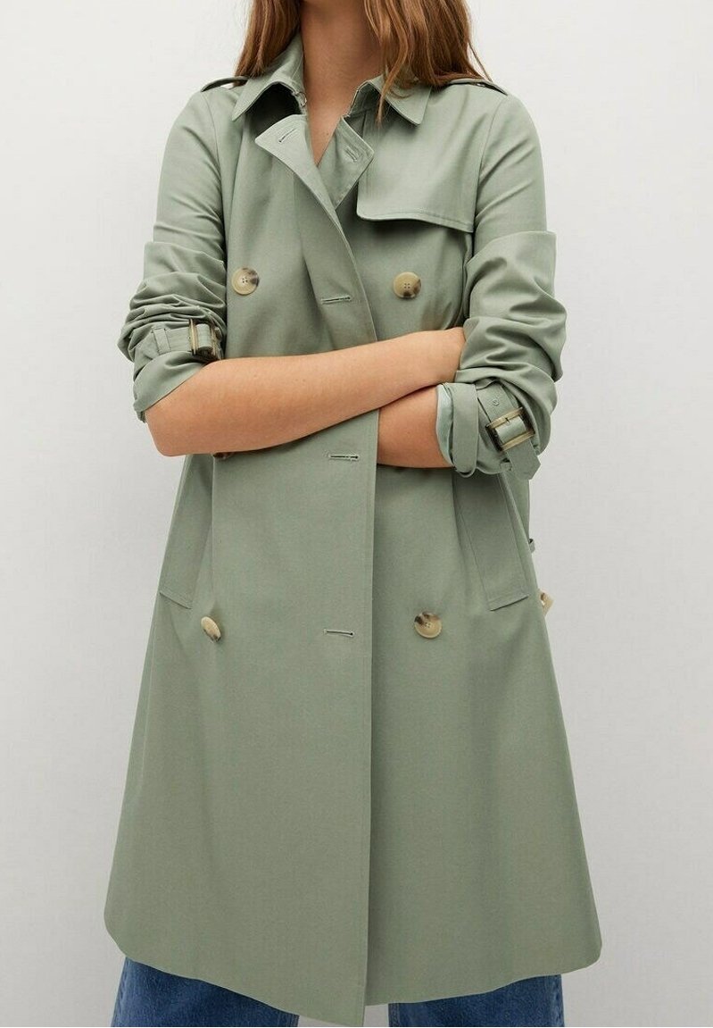 Femme portant un trench-coat vert clair à double boutonnage avec des manches retroussées et des boutons beiges, bras croisés, associé à un jean bleu.