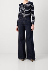 Marineblauwe gestreepte cardigan met knoopsluiting, gecombineerd met wijde donkerdenim jeans. De gestructureerde stof en gouden knoopaccenten zijn zichtbaar.
