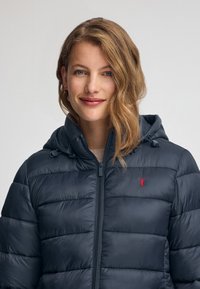 Marinblå puffjacka med dragkedja, huva och horisontella sömmar. Har en liten röd logotyp på bröstet och en slät yta.