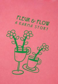 Rožinė marškinė su žalia grafika, vaizduojančia dvi puodelius su gėlėmis, tekstas "FLEUR & FLOW A KARMA STORY", švelni tekstūra, apvalus iškirpimas.