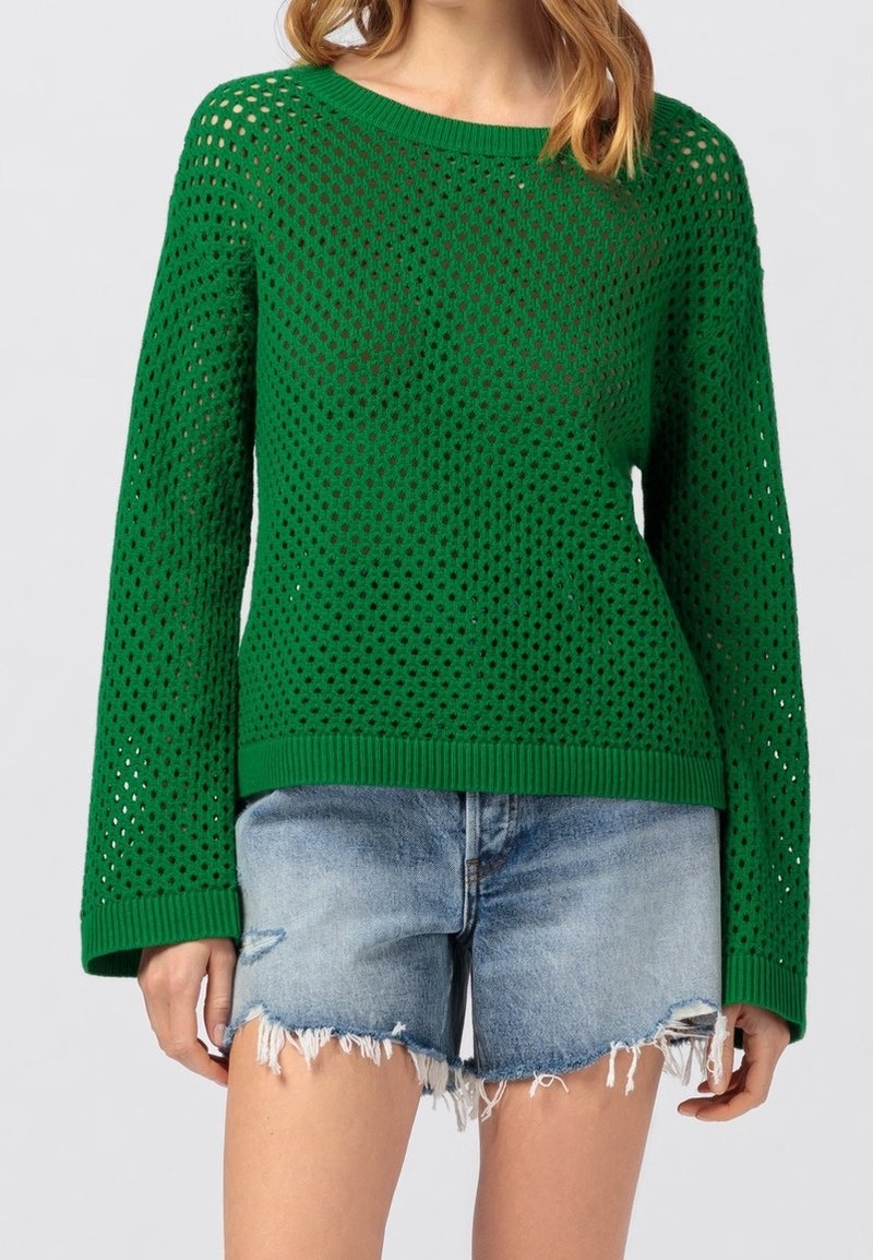 Grön öppenstickad tröja med långa ärmar, sammansatt med ljusblå, fransade denimshorts på en stående person.
