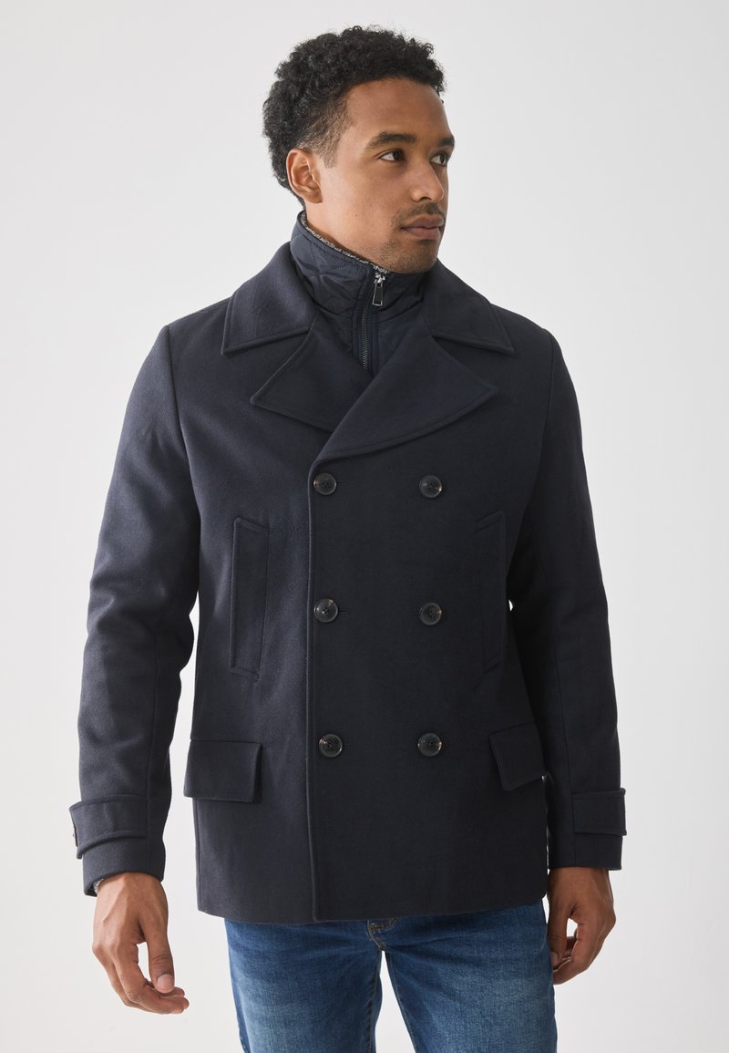 Manteau en laine marine doublé avec de grands revers, un col zippé et deux poches latérales ; texture en laine douce, avec boutons noirs.