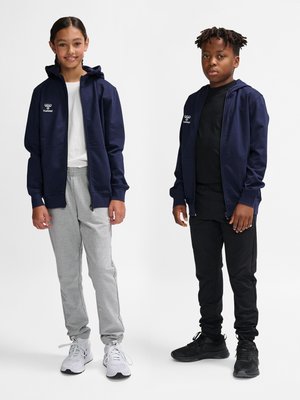 Dwoje dzieci stoi, ubrani w granatowe kurtki z kapturem; jedno w białej koszuli i szarych spodniach z białymi trampkami, drugie w czarnej koszuli i spodniach z czarnymi butami.