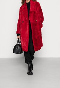 Nainen kävelee pitkään punaiseen tekoturkistakkiin pukeutuneena, mustat housut, mustat saappaat ja musta tikattu käsilaukku kädessään.