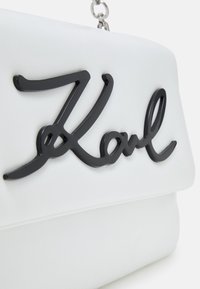 Borsa in finta pelle bianca con superficie texturizzata, caratterizzata da un notevole logo in corsivo nero "Karl". Include una tracolla in catena argentata.