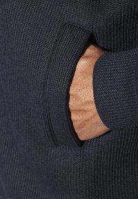 Marineblauer Strickpulloverstoff mit strukturiertem Muster, sichtbar ist eine Hand in einer seitlichen Tasche. Glatte, eng anliegende Bündchen.