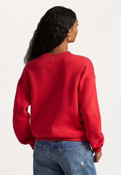 Felpa rossa con scollo rotondo, vestibilità rilassata, polsini e orlo a coste; abbinata a jeans blu strappati, per un look casual.
