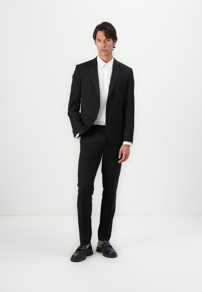 Costume noir ajusté, associé à une chemise blanche. Présente un léger éclat, une texture lisse et des chaussures en cuir noir.