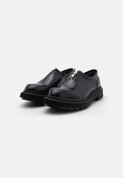 Black leather slip-on shoes with a high-gloss finish, featuring silver zippers and a thick textured rubber sole. Rounded toe design.