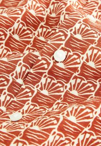 Tela de patrón rojo y crema con un diseño repetido de formas abstractas. Presenta botones blancos a lo largo del borde. Material de algodón con textura.