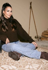 Bruine geribde trui, lichtblauwe jeans en donkerbruine schoenen. Zittend op een patroon tapijt, met houten accenten op de achtergrond.