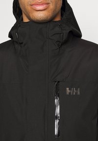 Chaqueta impermeable negra con cuello alto, cordón ajustable, bolsillo frontal con cremallera y un logotipo texturizado en el pecho. Acabado de tejido suave.