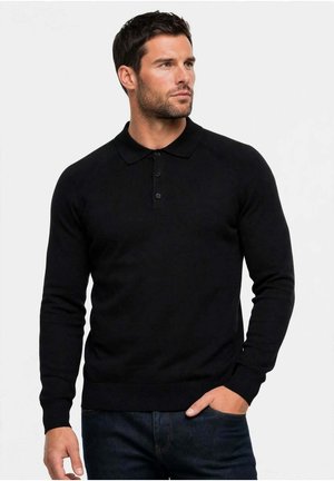 Hombre con pelo oscuro que lleva una camisa polo negra de manga larga y vaqueros oscuros, de pie con una mano en el bolsillo, mirando hacia un lado.