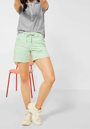 Femme portant une chemise rayée et un short vert menthe, se tenant les jambes croisées devant une chaise corail sur fond blanc.