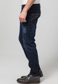 Mörkblå denimjeans med smal passform, med orange sömmar, klassisk femficksdesign och subtil blekning längs benen.