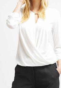 Blusa bianca a maniche lunghe con scollo a V e design frontale intrecciato. Presenta un tessuto morbido, una vestibilità comoda e un sottile accento dorato all'escollatura.