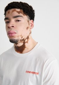 White cotton t-shirt featuring red "CONVERSE" text. The model has a closely cropped hairstyle and visible skin patterns on the face.