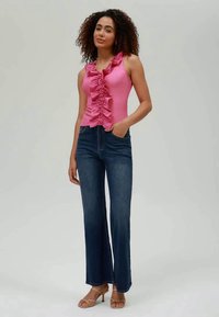 Pink sleeveless top with ruffled front, paired with dark blue flared jeans and beige high-heeled sandals. Smooth fabric, relaxed fit.