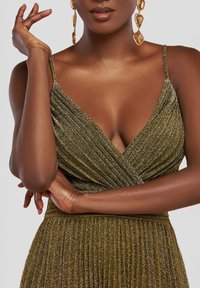 Metallisk olivengrøn kjole med plisseret design, V-hals og tynde stropper. Guld statementøreringe kompletterer outfittet.