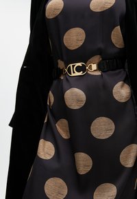 Schwarzes Kleid mit großen beigen Punkten, ausgestattet mit einem Gürtel mit goldener Schnalle und Logo. Weicher Stofftextur, begleitet von einem schwarzen Mantel.