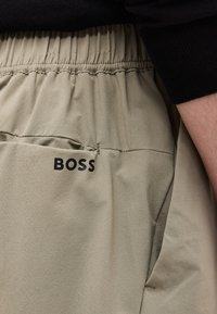 Pantalon beige à taille élastique avec poche affichant le logo noir "BOSS", porté avec un haut noir à manches longues, bras partiellement visible.