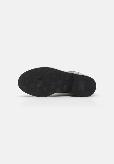 Suela de zapato de goma negra con patrón texturizado en relieve y detalle de marca. Forma redondeada, diseño plano y apariencia minimalista.