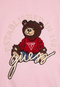 Sudadera rosa con un oso bordado que lleva un suéter rojo y pantalones cortos azules, con el logo de "Guess" y el texto "Establecido en 1981".