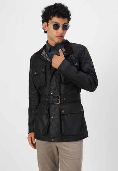 Veste noire imperméable avec quatre poches avant, une taille ceinturée et un col texturé marron. Modèle portant des lunettes de soleil et une chemise à carreaux en dessous.