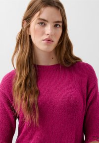 Maglione fucsia lavorato a maglia con texture a coste, scollatura rotonda e vestibilità ampia. Il tessuto ha un aspetto morbido e voluminoso, che ne enfatizza il calore.