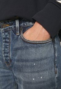 Jeans i denim med mörk tvätt, med lätt blekning, sömnadsdetaljer och vita färgfläckar på vänster lår. Ficka synlig.