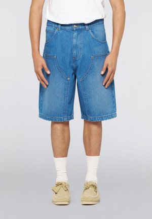 Persona che indossa pantaloncini di jeans blu fino al ginocchio, calzini bianchi, scarpe beige e una camicia bianca, in piedi davanti a uno sfondo semplice.