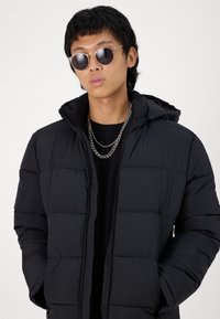 Svart puffer-jakke med hette, med quiltemønster og glidelås. Modellen har på seg sølvkjeder og runde solbriller.