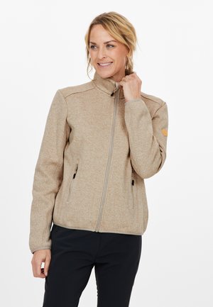 Veste zippée beige avec un col haut, deux poches latérales et un tissu texturé. Présente une fermeture éclair contrastante et un patch logo sur la manche.