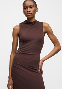 Γυναίκα με πλεξούδες, που φοράει ένα αμάνικο, στενό, σκούρο καφέ φόρεμα με ψηλό λαιμό, στέκεται με το ένα χέρι στη μέση, ουδέτερο φόντο.